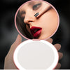 A hand holds an open personalized LED makeup mirror showing a close-up of a woman's eye and lip makeup.