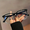 A hand is holding a pair of black-framed, clear-lensed minimalist square reading glasses against a light background.