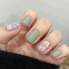 A hand displays a set of press-on acrylic flower nails with a pale green base and iridescent glitter accents.