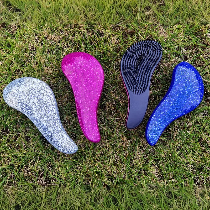 Four brightly colored detangling hair brushes—silver, pink, black, and blue—lie on green grass.