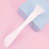 A white silicone facial mask brush with an integrated scraper resting on a light blue square box.