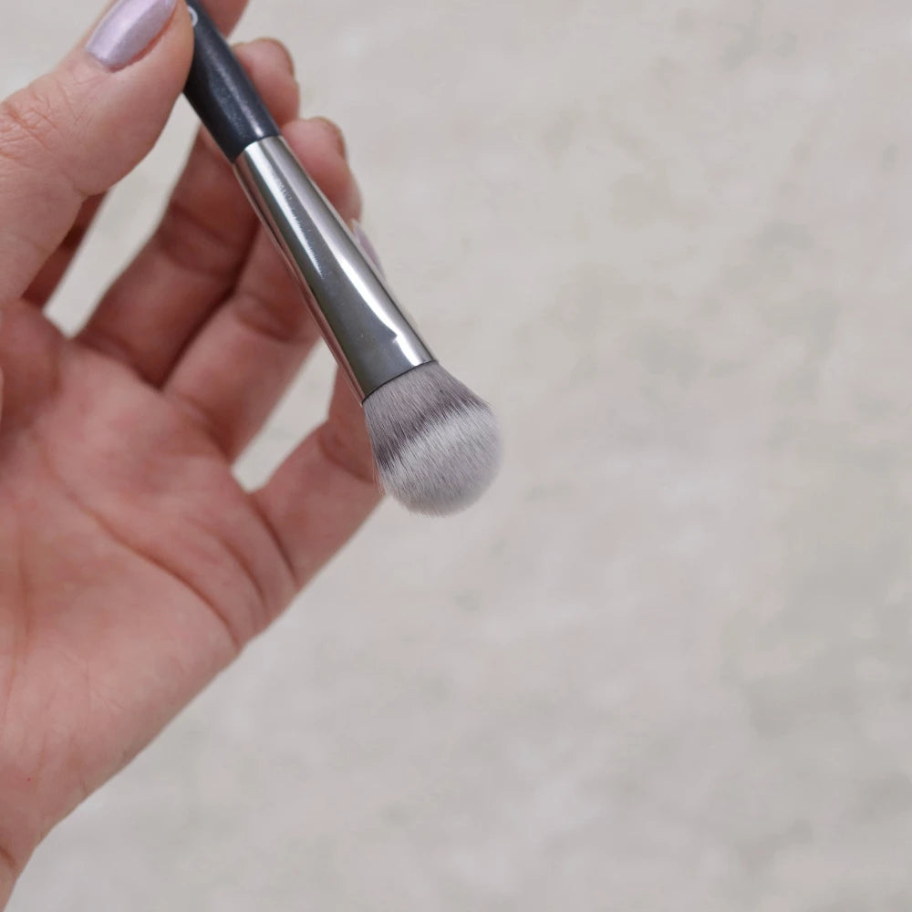 A hand holds a precision concealer brush with a silver ferrule and soft, dense bristles.