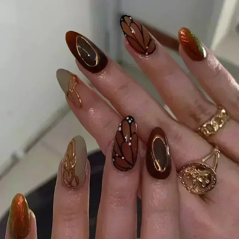 A close-up of a hand displaying 24-piece almond press-on nails featuring 3D floral and butterfly designs in various earthy to