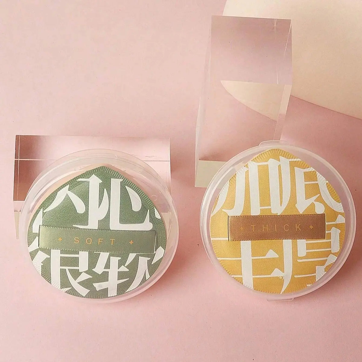 A pair of dual-sided makeup sponge puffs in green and yellow packaging are shown on a pink surface.