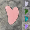Masajeador facial de cera en forma de corazón rosa, mostrado sobre tela gris texturizada con otros corazones de colores.