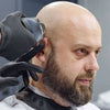 A man with a beard is being shaved by a person wearing black gloves using a precision electric shaver.