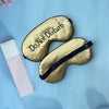 A gold silk sleep mask embroidered with the text Do Not Disturb is shown on a light blue surface.