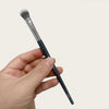A hand holds a black precision concealer brush with a silver ferrule and a white synthetic tip.