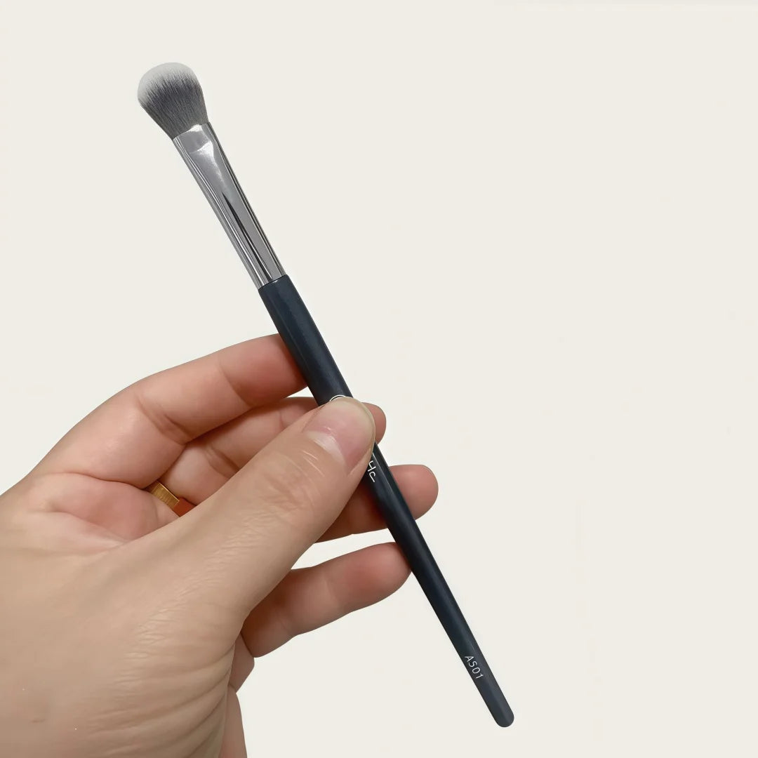 A hand holds a black precision concealer brush with a silver ferrule and a white synthetic tip.