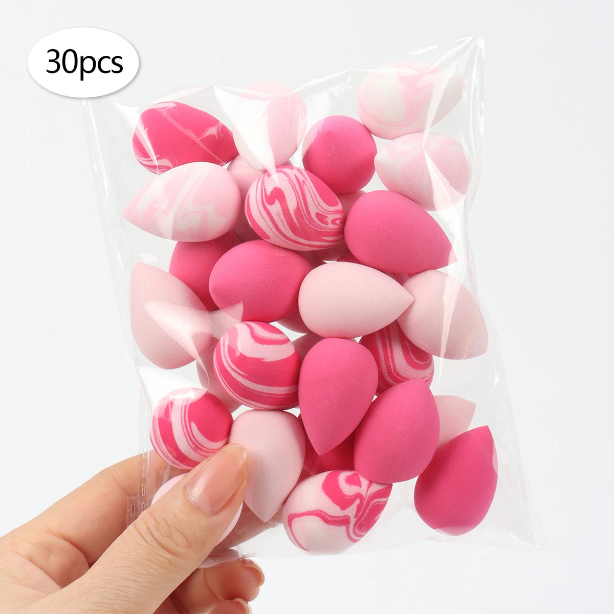 A hand holds a clear plastic bag containing 30 reusable pink and white expandable makeup sponges.