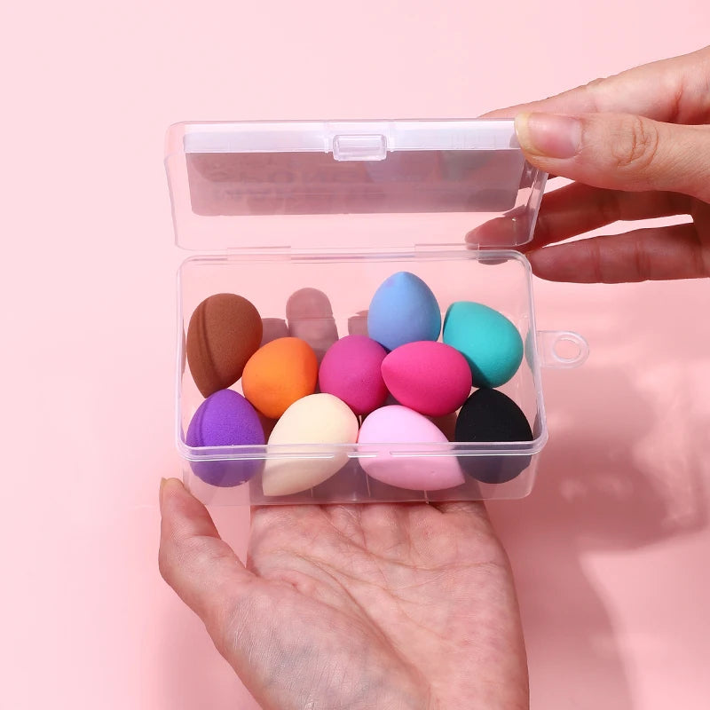 A hand holds an open clear plastic container filled with colorful makeup sponge puffs.