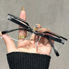 Una mano sostiene unas gafas de lectura cuadradas negras anti-luz azul con monturas transparentes y detalles dorados.