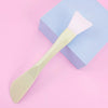 A pale yellow silicone facial mask brush with a white head rests on a light purple box.