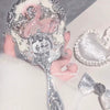 A silver-toned makeup mirror set with an ornate frame is being held by a hand, displayed on a white surface.