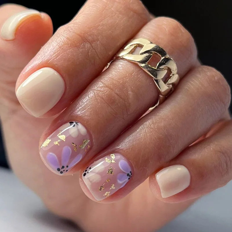 A hand models blue floral press-on false nails with gold accents and a gold link ring.