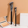 A set of two black professional makeup brushes standing upright on a beige background next to an open makeup palette.
