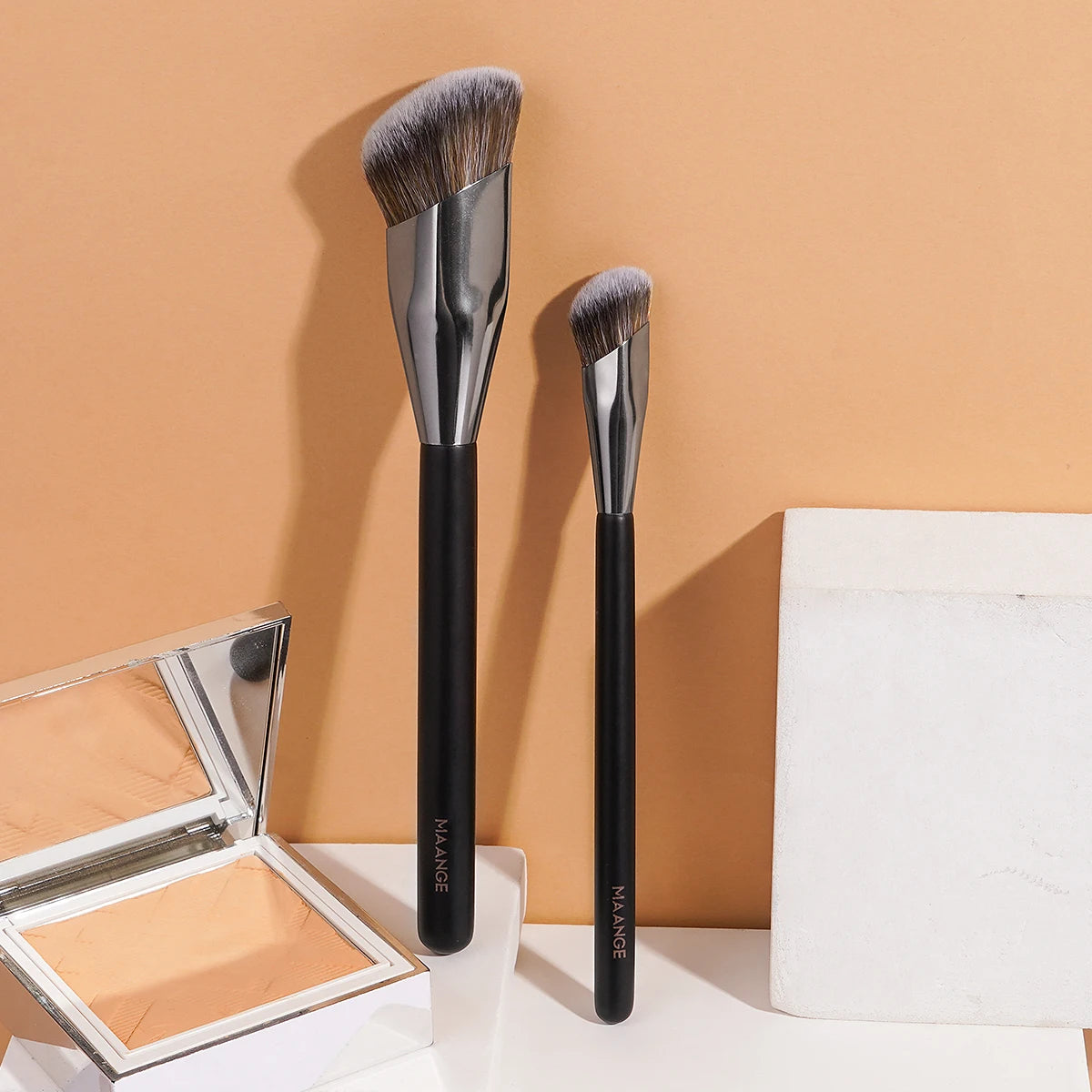A set of two black professional makeup brushes standing upright on a beige background next to an open makeup palette.