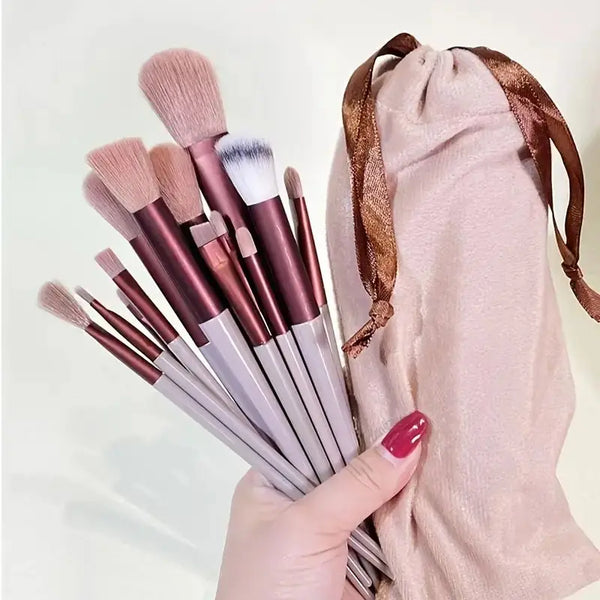 A hand holds a set of thirteen professional makeup brushes with various shades and a pink fabric pouch.