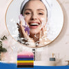 Une femme applique une éponge exfoliante violette sur son visage, enveloppée dans une serviette blanche.