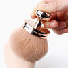 Une main tient une grande brosse de maquillage ronde et douce, de couleur marron avec un manche doré.