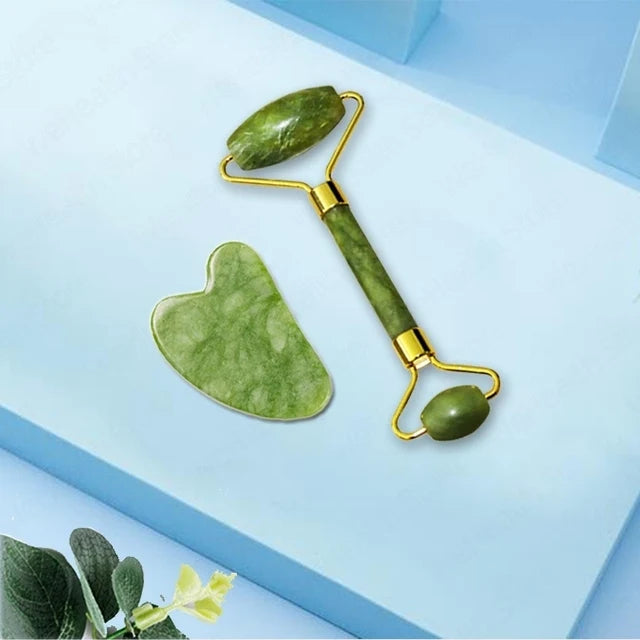 A jade gua sha facial massager tool with a green stone and gold handle resting on a light blue surface.