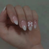 A hand displaying almond press-on nails with a sheer pink gradient and a delicate white bow design.