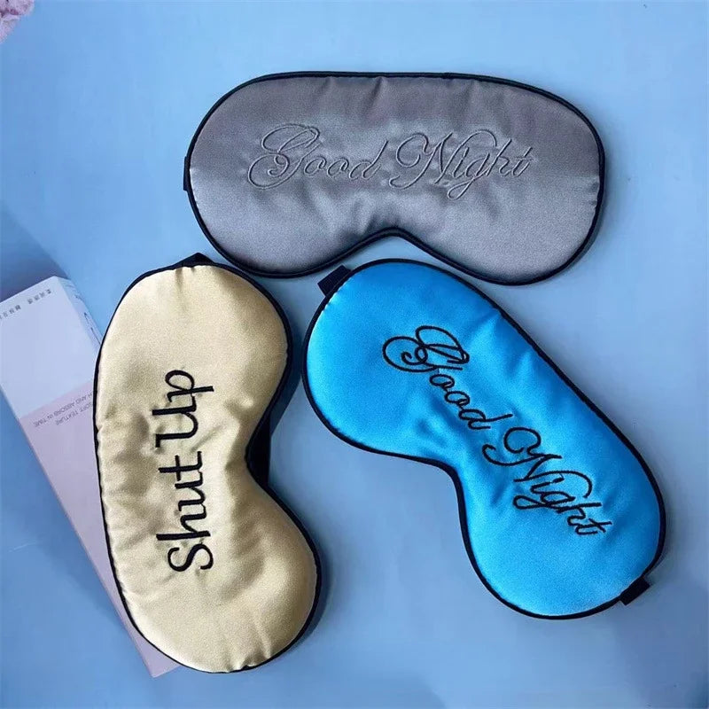 Three embroidered silk sleep masks are displayed on a light blue surface, featuring 'Good Night' and 'Shut Up' designs.