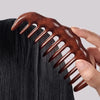 A hand holds a brown wooden wide-tooth detangling comb against dark hair.