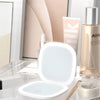 A compact LED makeup mirror with a white frame and a clear magnifying lens is shown on a glass surface.