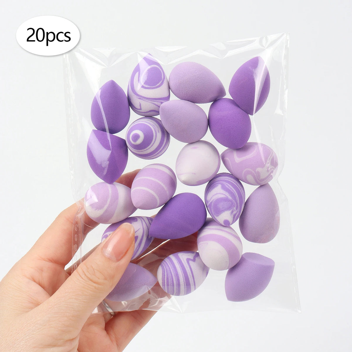 A hand holds a clear plastic bag containing 20 reusable expandable makeup sponges in various shades of purple and lilac.