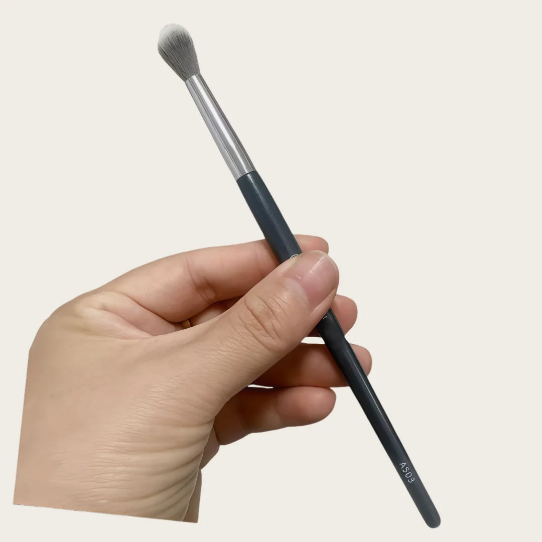 A hand holds a black precision concealer brush with a silver ferrule and a tapered tip.