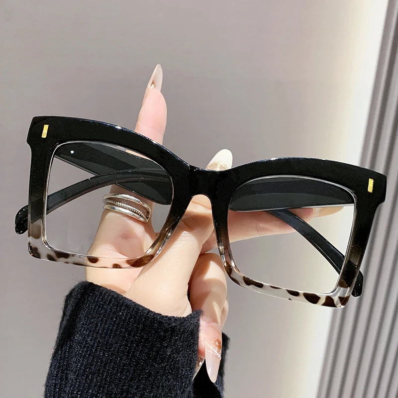 A hand holds a pair of black square frame reading glasses with tortoiseshell accents against a light background.