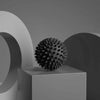 A dark gray, spiky massage ball rests on a light gray rectangular block against a muted background.