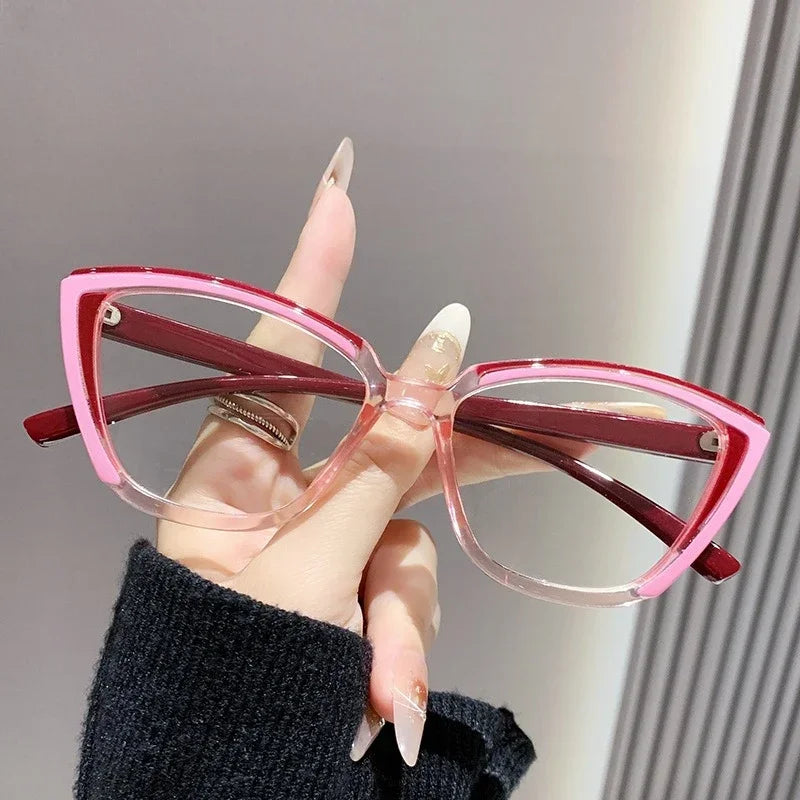 A hand holds a pair of pink and maroon cat eye reading glasses against a light gray background.