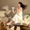A woman is using a professional high-speed hair dryer indoors while sitting on a sofa.