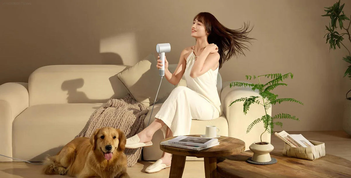 A woman is using a professional high-speed hair dryer indoors while sitting on a sofa.