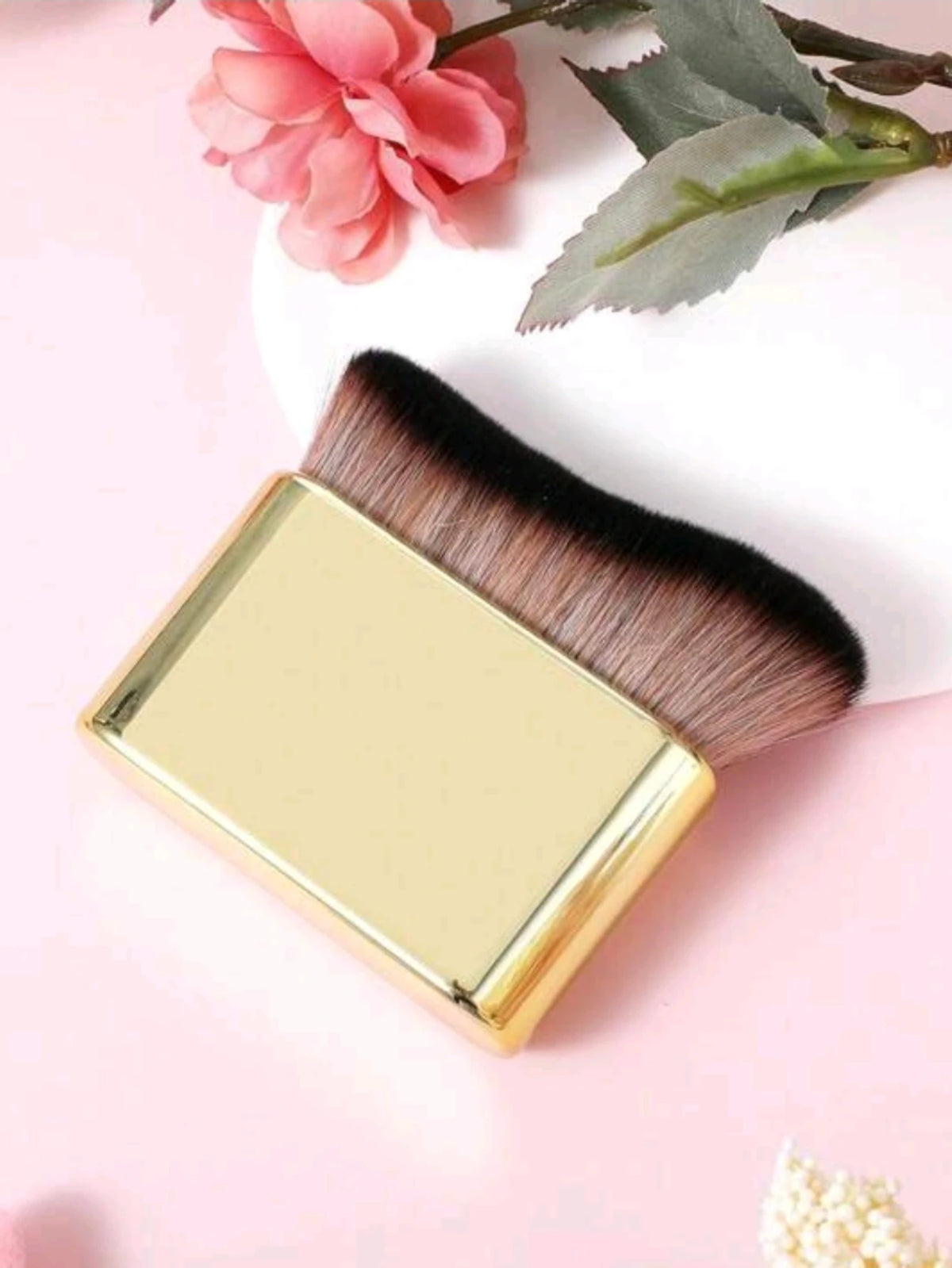 Un set de pinceaux de maquillage de couleur or avec des poils foncés est exposé sur un fond rose à côté d'une fleur.