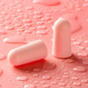 Pink memory foam earplugs resting on a textured pink surface with water droplets.