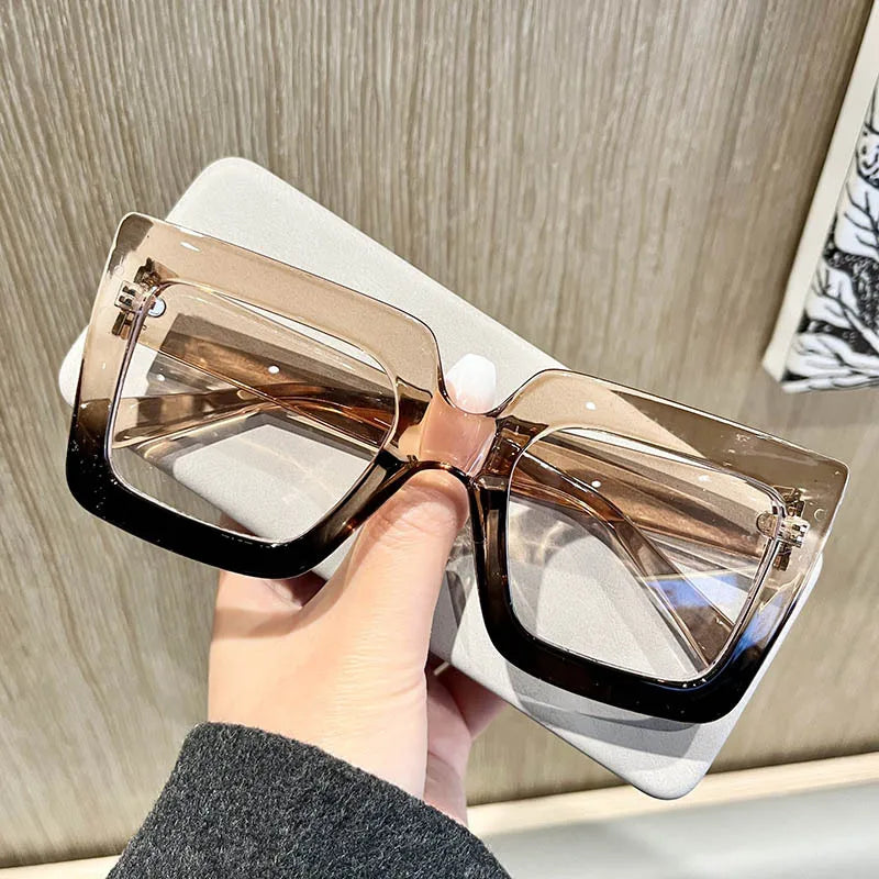 A hand holds a pair of classic square eyeglasses with clear brown lenses against a light background.