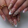 A close-up of a hand displaying 24-piece almond press-on nails featuring gold and pearl floral and butterfly designs.