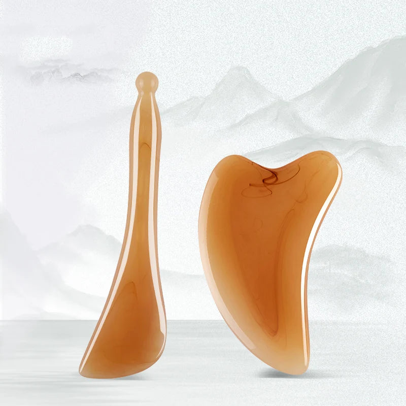 A set of two amber-colored facial massage and lifting tools displayed against a white background with mountains.