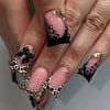 A close-up of a hand displaying nude pink press-on nails with black leopard print and gold 3D charms.