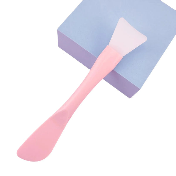 A pink silicone facial mask brush with an integrated scraper resting on a light blue square surface.