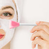 Una mano aplica un cepillo de mascarilla facial de silicona rosa sobre una mascarilla facial blanca en el rostro de una perso