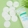 A collection of white disposable cotton pads for makeup removal and face cleaning are displayed on a green surface.