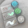 A collection of iridescent glitter powders in shades of blue, green, and silver displayed on a sparkly surface.