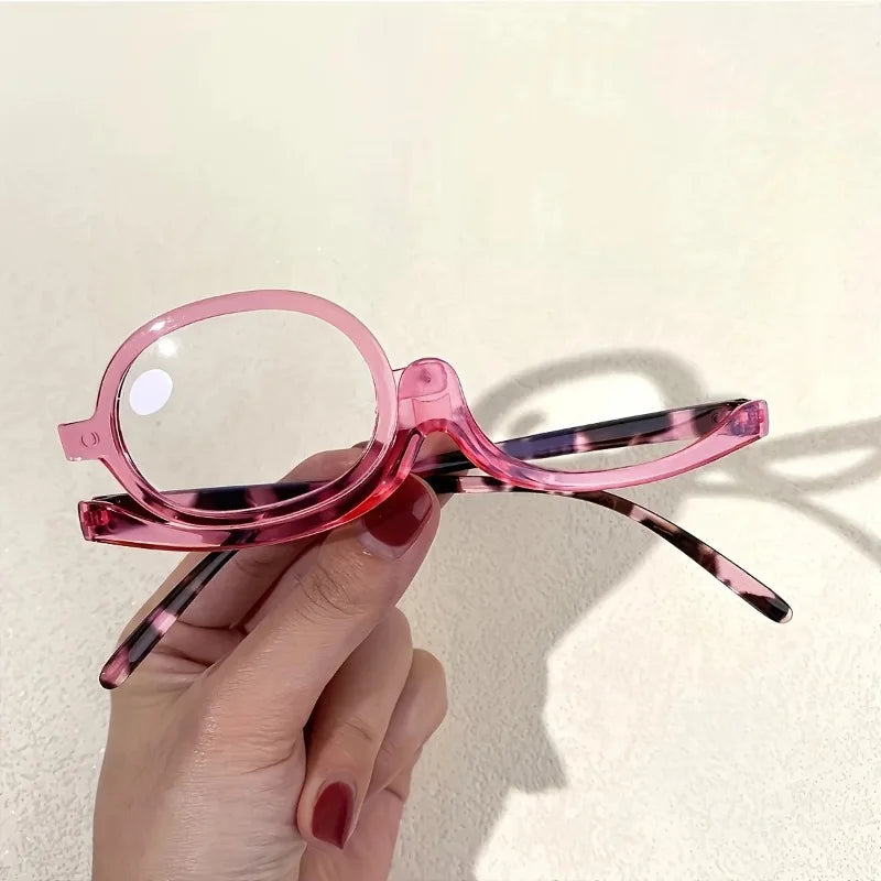 A hand holds a pair of pink and purple marbled magnifying reading glasses against a light background.