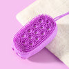 A purple silicone body brush with numerous bristles rests on a white towel against a pink background.