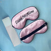 A pink silk sleep mask embroidered with the text Leave Me Alone is displayed on a light blue background.