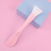 A pink silicone facial mask brush with an integrated scraper rests on a light blue square box.